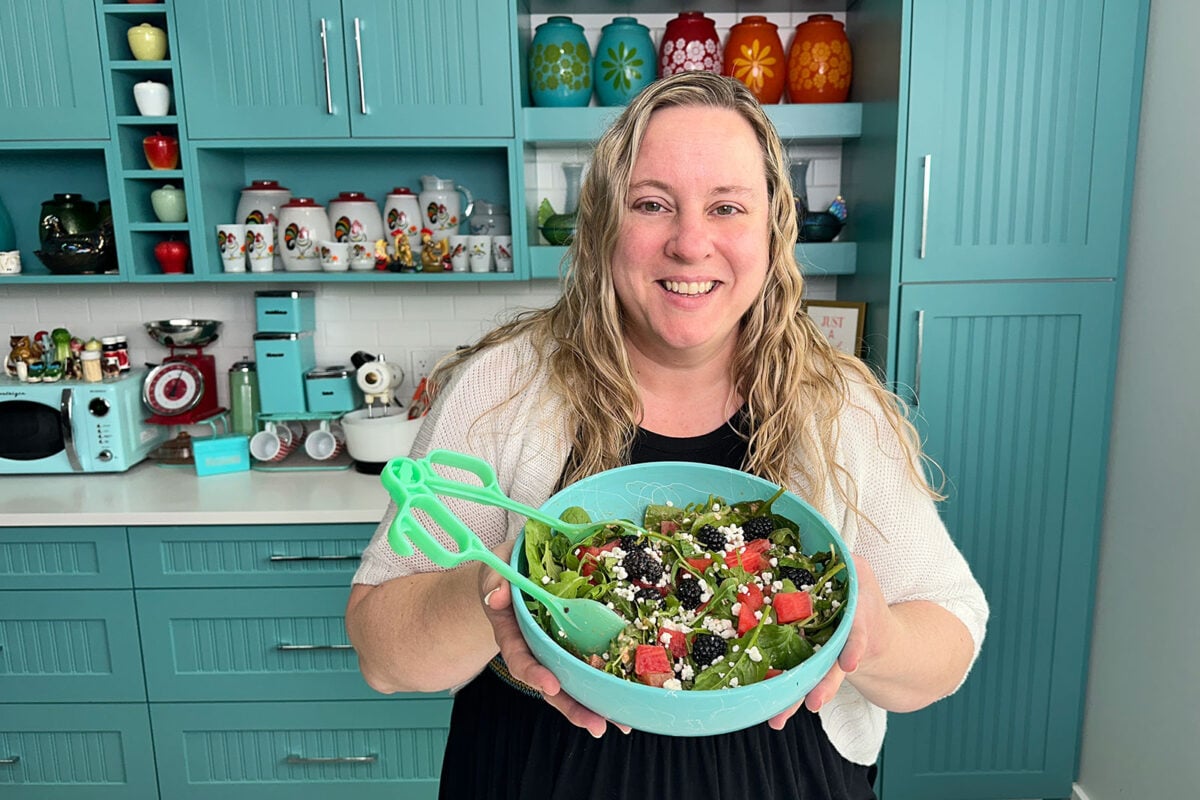 Karlynn holing a bowl of watermelon salad with feta and mint in a green bowl with green tongs.